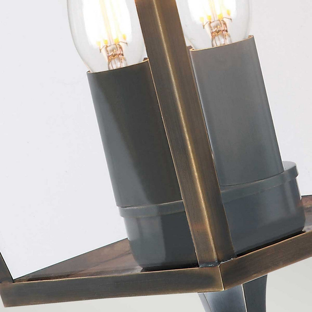 Close-up of a Mansion 3 Light Outdoor Wall Light in aged brass, highlighting its modern design with two exposed bulbs inside a geometric frame accented by black details. The illuminated bulbs contrast against a light background, exemplifying exquisite British craftsmanship.