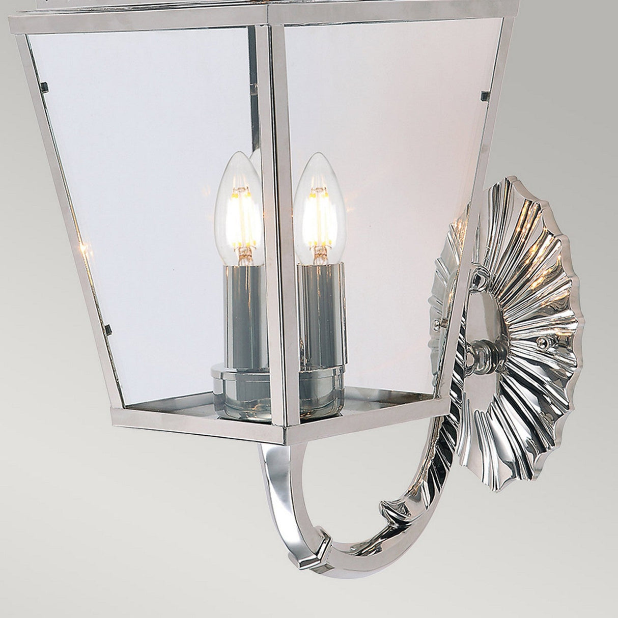The Mansion 3 Light Outdoor Wall Light in polished nickel boasts a lantern-inspired design with a geometric glass enclosure housing two visible candle-shaped bulbs, and its base is adorned with a decorative, fan-like detail.