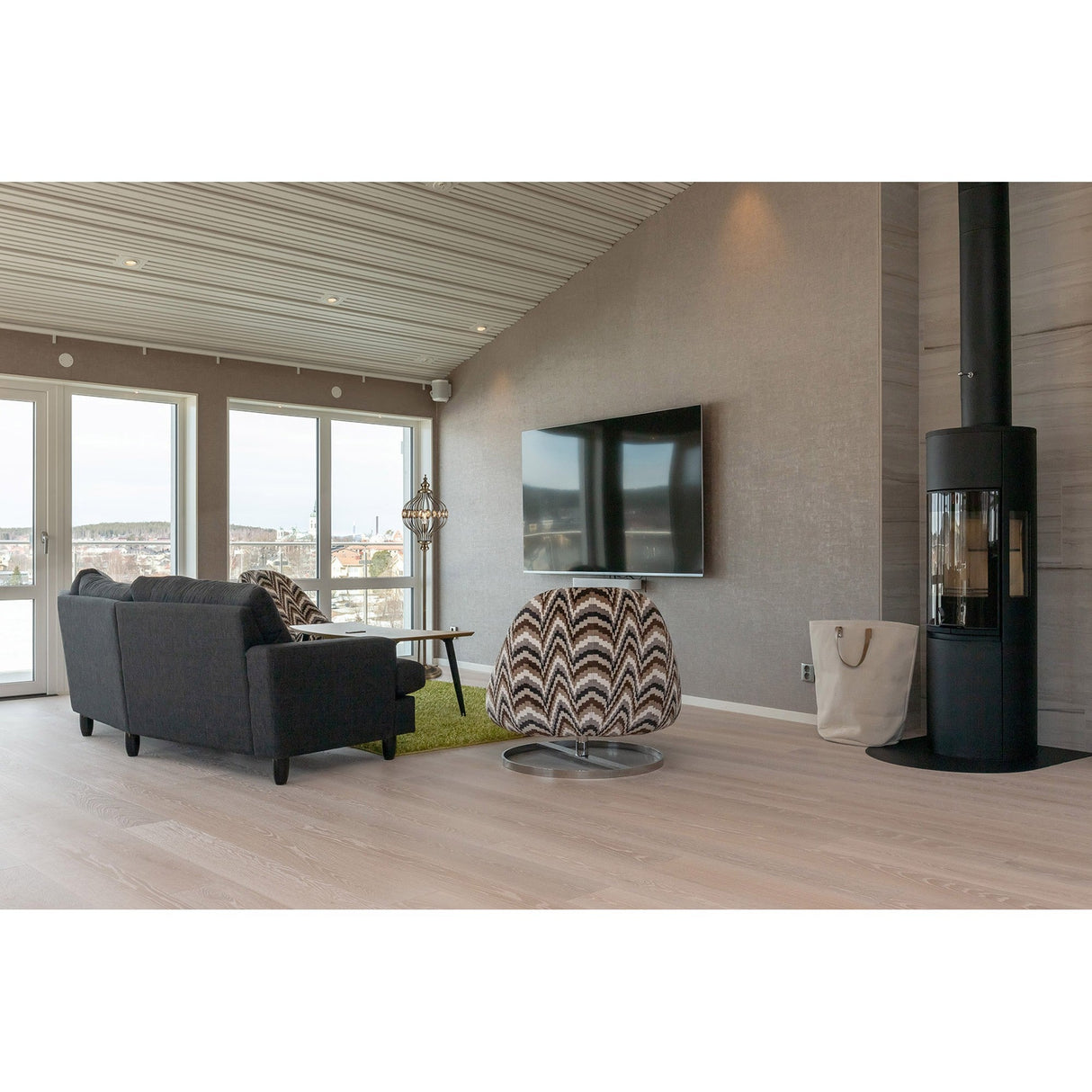 Modern living room with light wood floors, a gray sofa, patterned swivel chair, Marisa 1 Light Crystal Floor Lamp in antique brass, wall-mounted TV, black cylindrical wood-burning stove, and large windows overlooking the water.