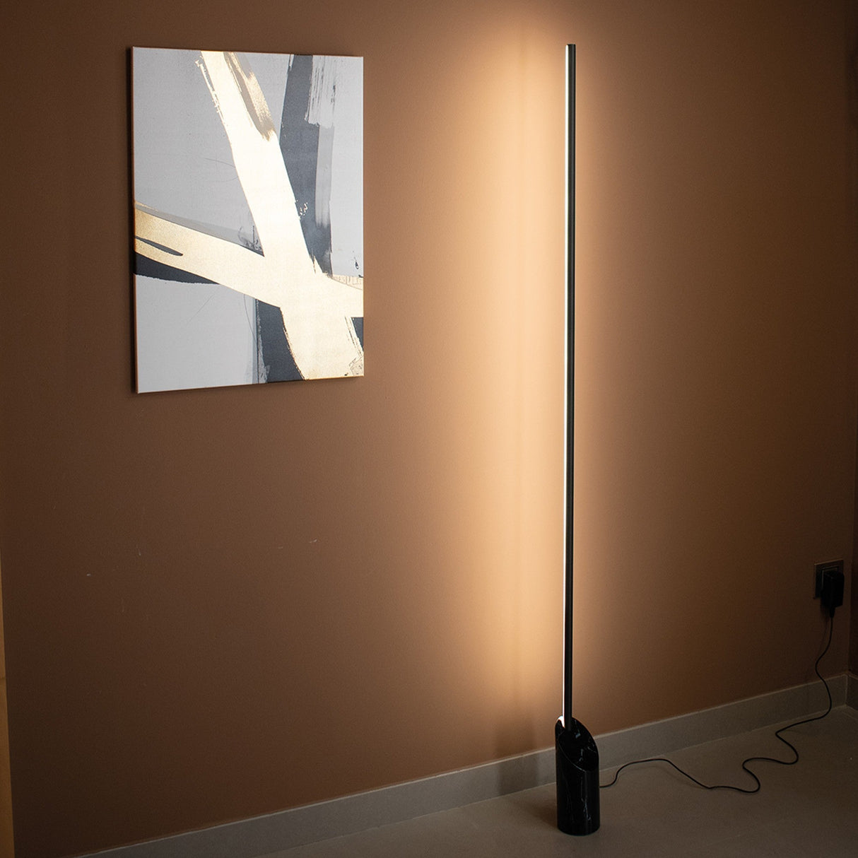 A modern, minimalist room with a brown wall features abstract wall art and the Marmol LED Floor Lamp 28W 3000K in Black Marble & Black, emitting warm light, its cord plugged into a nearby outlet.