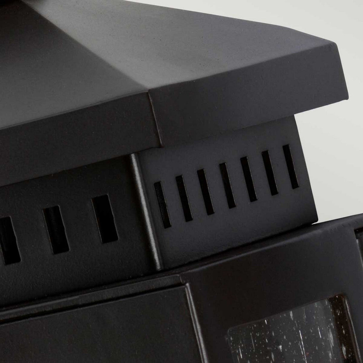 Close-up of the Matlock 1 Light Outdoor Wall Light in an old bronze finish, showcasing a black metal ventilated structure with a rectangular design and slotted vents. The matte surface exudes rustic elegance, while the transparent section reflects light beautifully. With its IP44 rating, it’s perfect as an outdoor wall light.