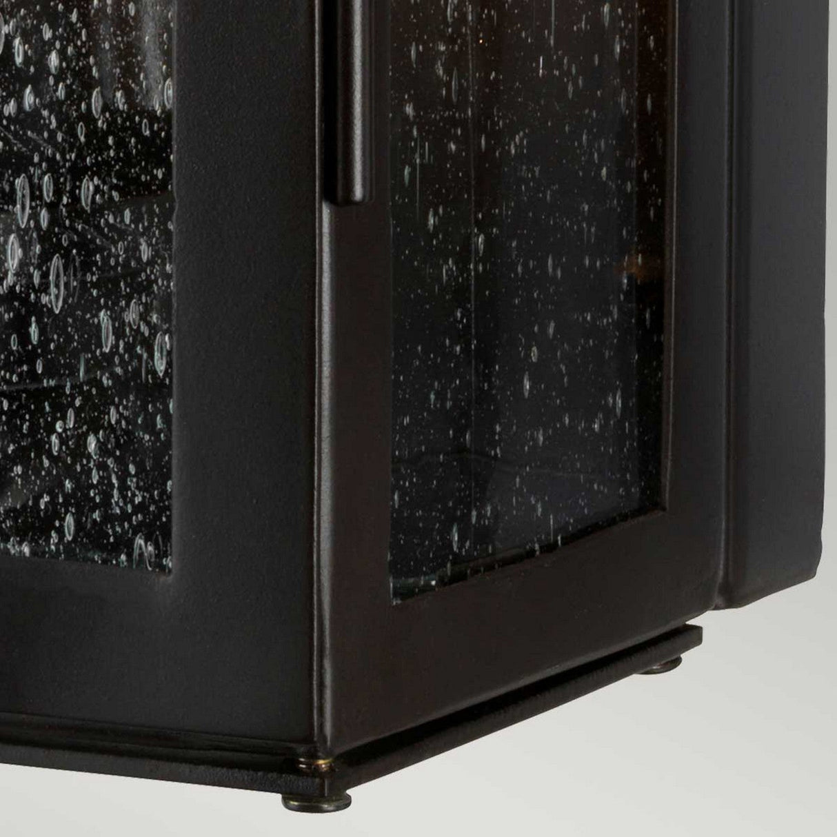 A close-up of the Matlock 1 Light Outdoor Wall Light in an old bronze finish showcases its rustic elegance, featuring black metal with bubbled glass panels. The design includes visible hinges and screws, highlighting its angular structure suited for outdoor use. With an IP44 rating, this lantern is set against a neutral background.