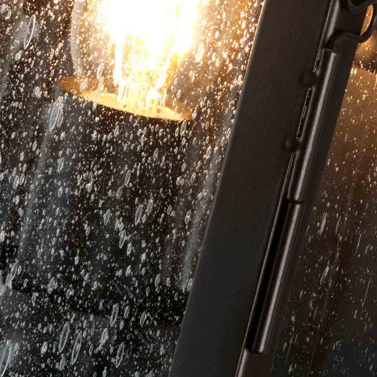 A close-up shot captures the glow of the Matlock 1 Light Outdoor Wall Light in Old Bronze as it shines behind a rain-speckled window. Droplets scatter across the glass, and the light casts an illumination that evokes rustic elegance and warmth, even against the wet exterior.