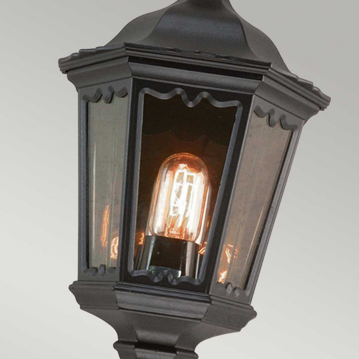 A close-up of the Medstead 1 Light Outdoor Wall Light With PIR - Black showcases its vintage-style charm with an illuminated bulb inside. This hexagonal lantern, featuring a sleek black finish metal frame and clear glass panels, stands out beautifully against a soft gradient of light gray background.