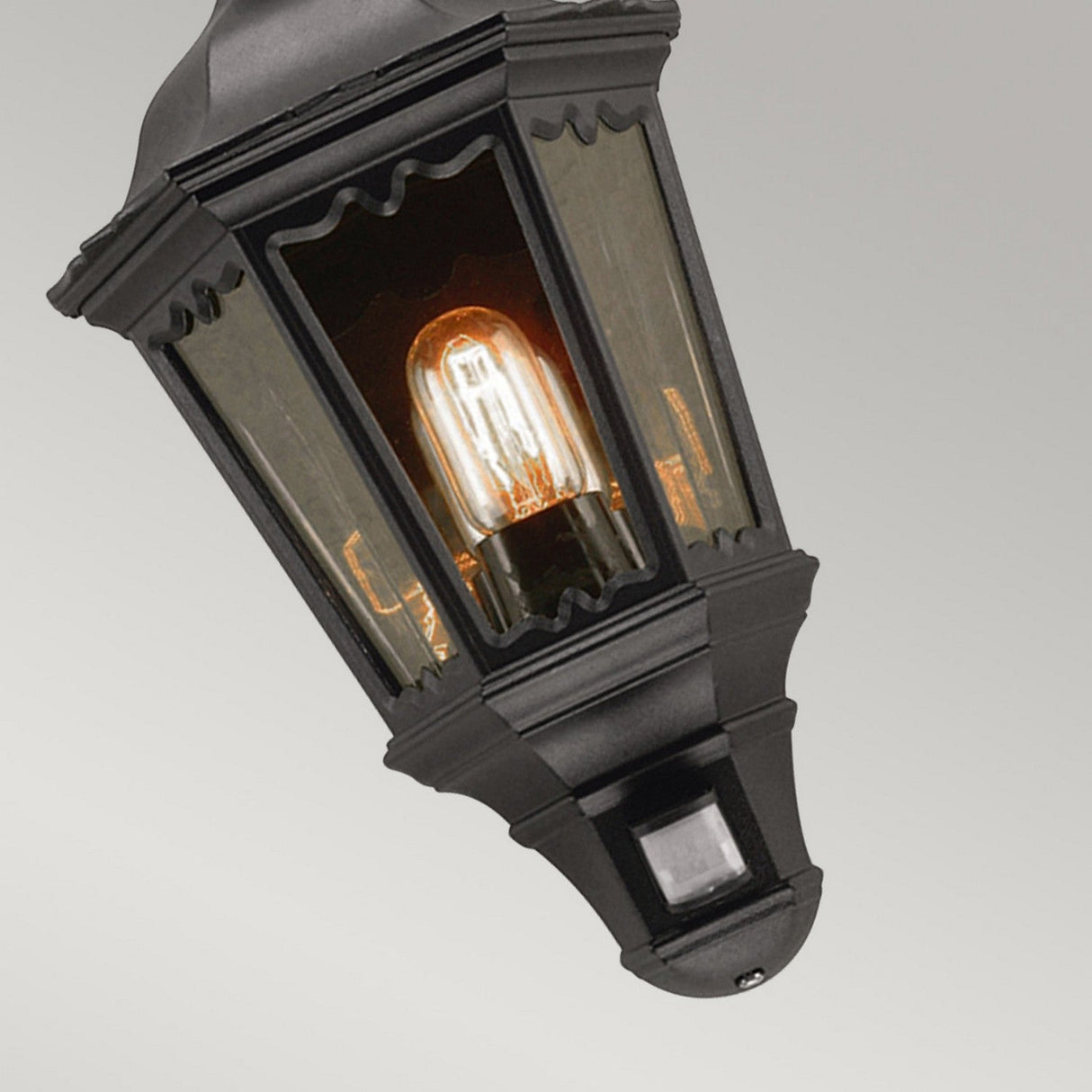 The Medstead 1 Light Outdoor Wall Light With PIR - Black showcases a vintage-style design with a glowing bulb encased in a black metal frame and clear glass panels. Elegantly mounted on a wall bracket, it is IP44 rated for weather resistance.