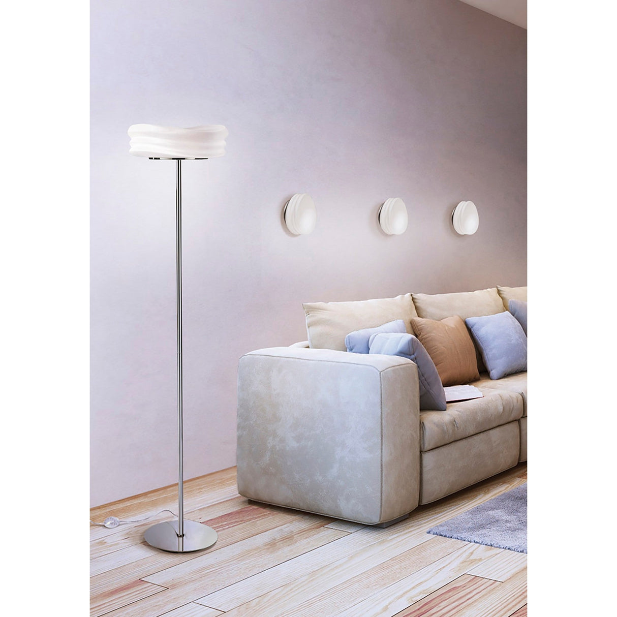A modern living room with a beige sectional, blue and tan cushions, and the Mediterraneo 2 Light Glass Floor Lamp in polished chrome with a white shade. Three round wall decorations on a light pink wall and pale wooden flooring complete the space.