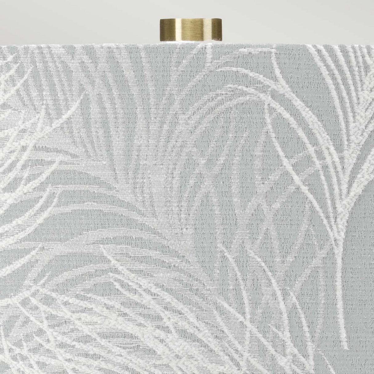 The Milo 1 Light Table Lamp features a light gray textured lampshade with an intricate white leaf pattern, elegantly perched on a ceramic base, and is topped off with an aged brass finial.