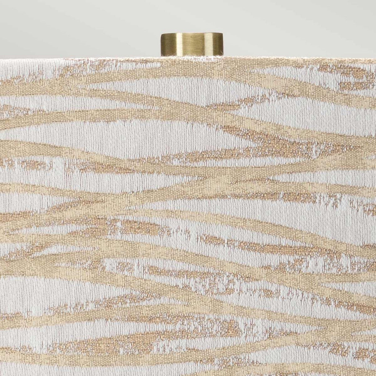 A detailed view of the Milo 1 Light Table Lamp reveals a decorative surface featuring an abstract pattern of wavy beige and white lines. The intricate texture shimmers subtly, paired with a partially visible cylindrical cap crafted from aged brass, accentuating its role as an elegant table lamp.