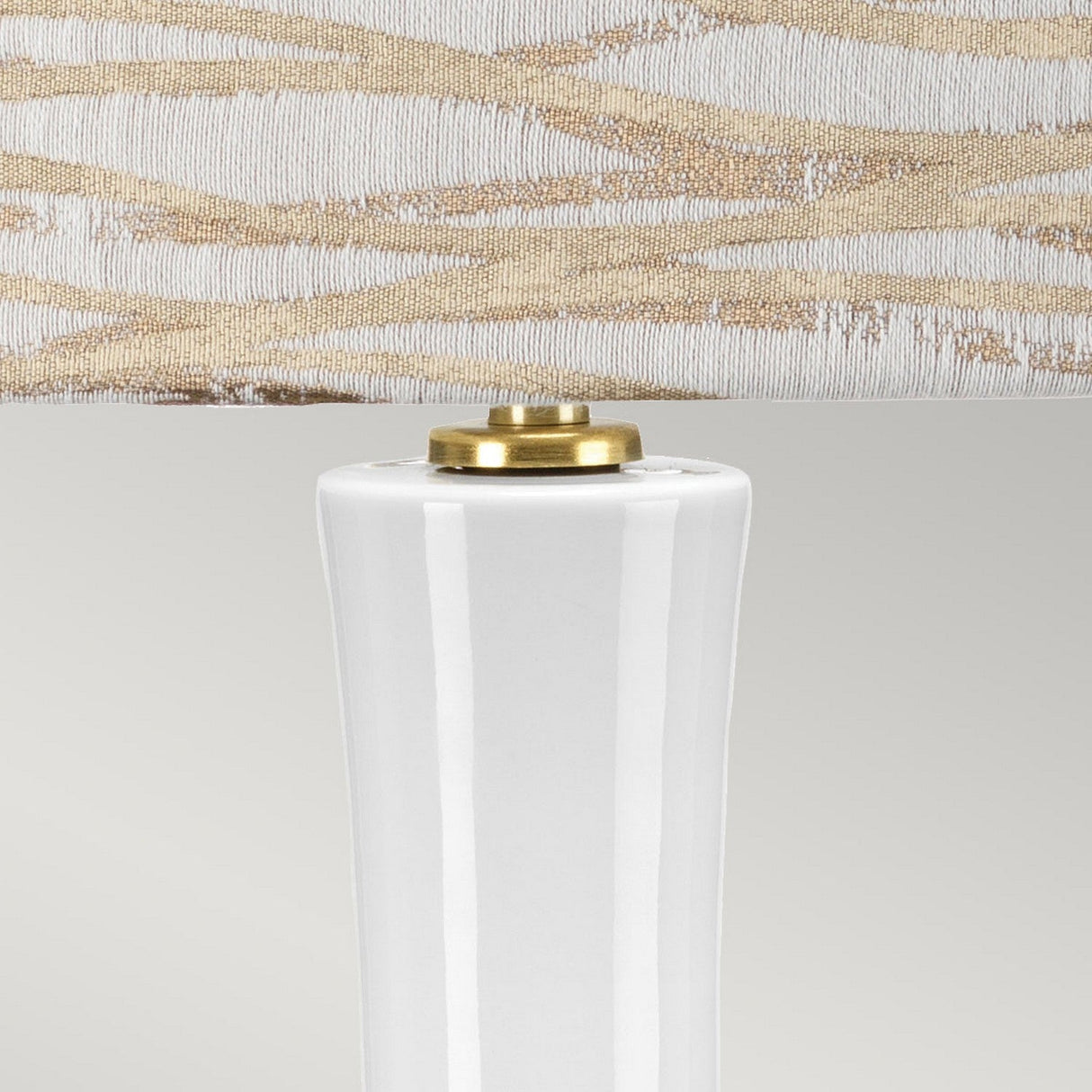 Close-up of the Milo 1 Light Table Lamp featuring a ceramic base with aged brass fittings, topped with a lampshade. The lampshade displays a textured pattern of beige wavy lines against a white background, exuding a modern and elegant aesthetic.