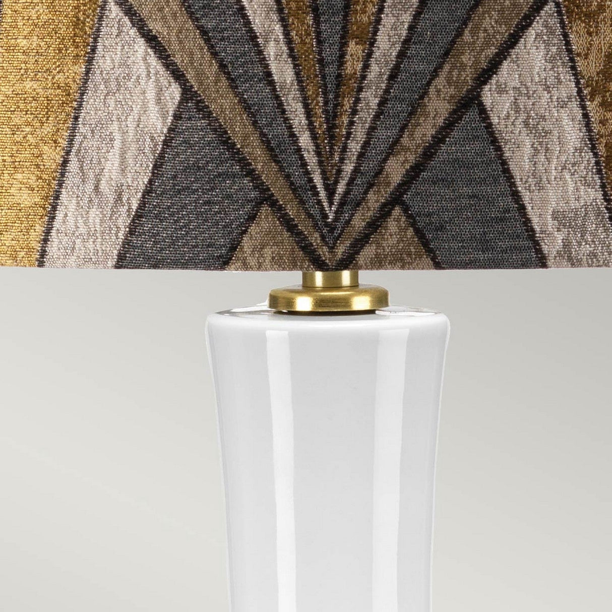 The Milo 1 Light Table Lamp in Aged Brass features a white shade with geometric patterns in brown, gold, and gray tones. This elegant lamp connects the decorative shade to a ceramic base with a sophisticated fixture of aged brass, all beautifully displayed against a plain background.