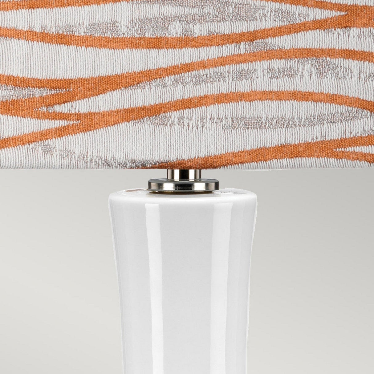 A close-up of the Milo 1 Light Table Lamp showcases its white ceramic base paired with an orange shade adorned with beige wavy patterns, accentuating the polished nickel detailing at both the top of the base and the lower part of its stylish design.