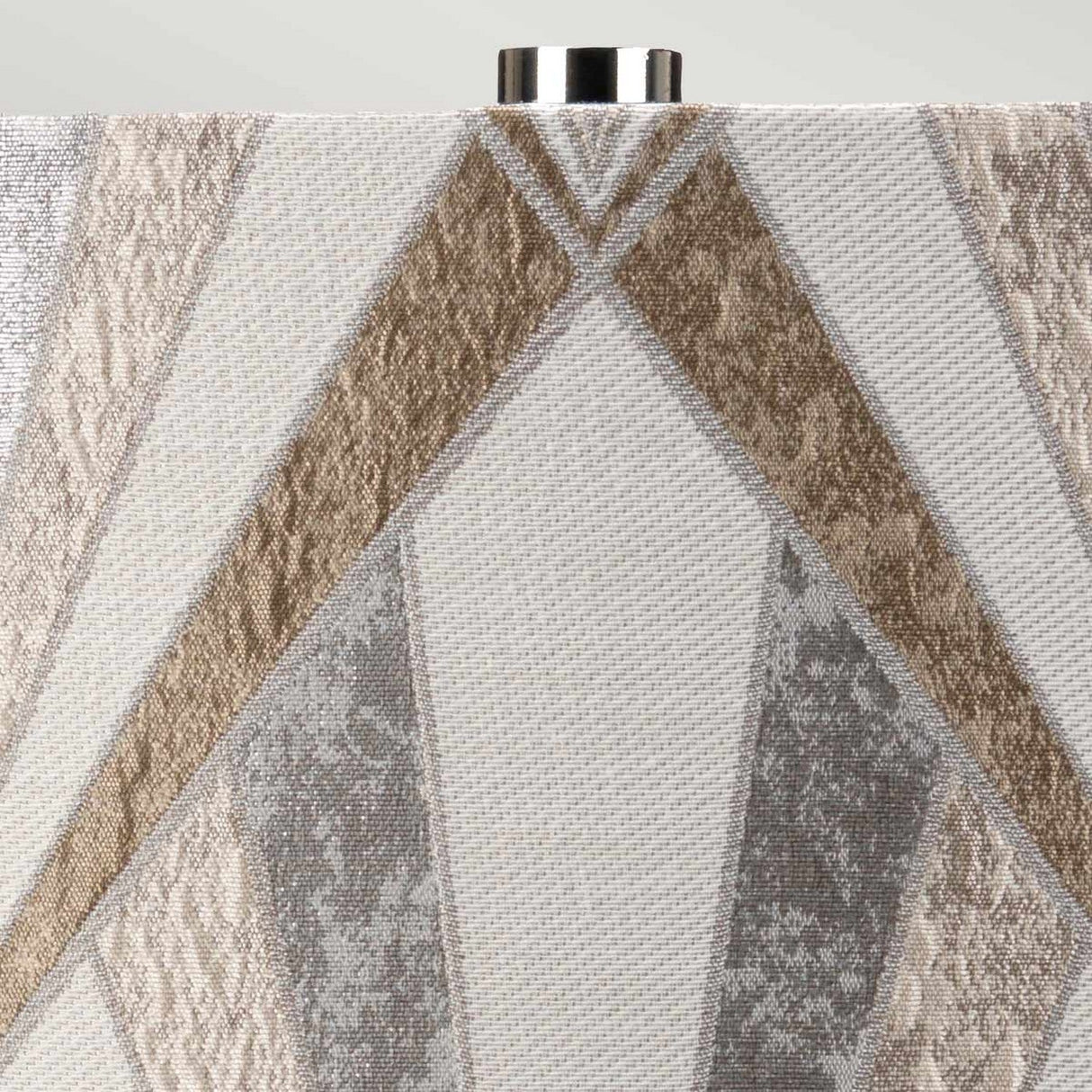A close-up of the fabric reveals an abstract geometric pattern in shades of beige, gray, and white, highlighted by metallic accents. At the top, a silver metal piece is partially visible, suggesting it could be part of the Milo 1 Light Table Lamp with a polished nickel finish.