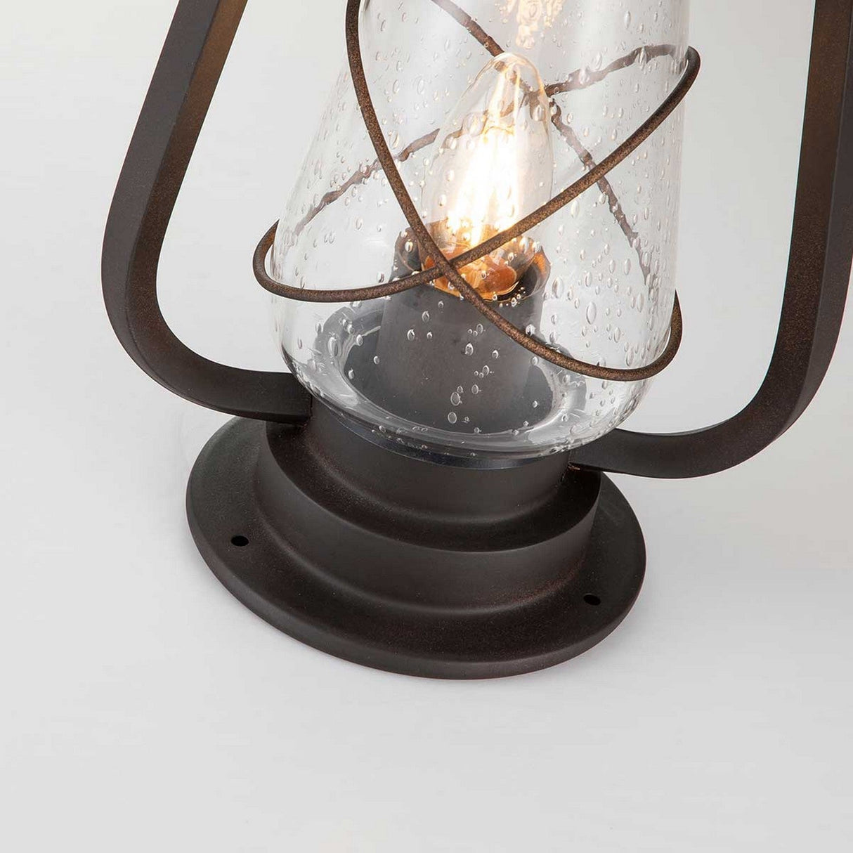 The Miners 1 Light Pedestal Light in Old Bronze showcases a rustic outdoor design akin to an antique lantern. It boasts a clear glass cover with small bubbles and houses an Edison-style bulb. Its decorative dark metal frame enhances its vintage charm.