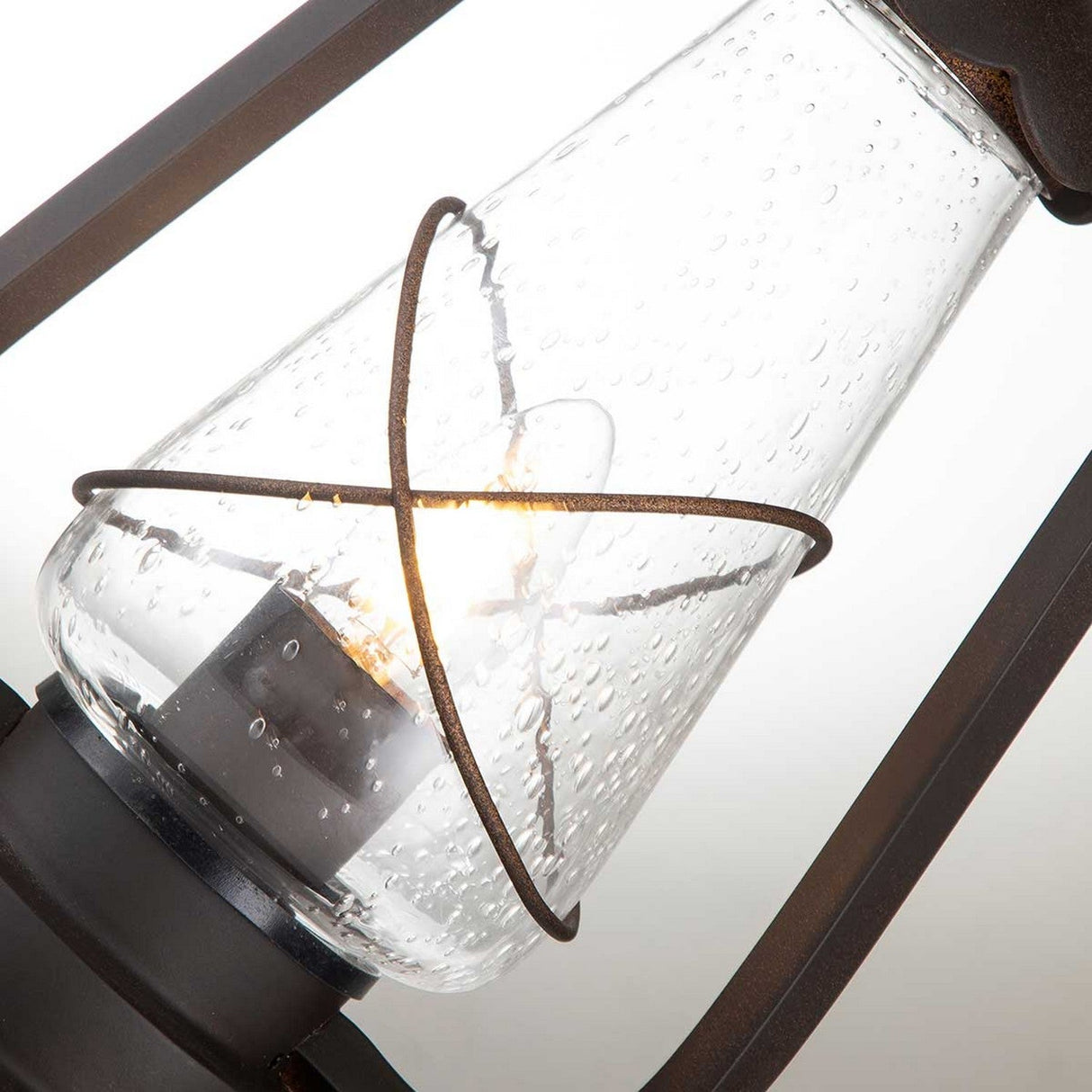 Close-up of the Miners 1 Light Pedestal Light - Old Bronze. The design showcases a clear, textured glass shade with a small glowing bulb inside, encased in a dark metal frame. The bubbled texture in the glass enhances its rustic charm, making it an ideal choice for an outdoor setting.