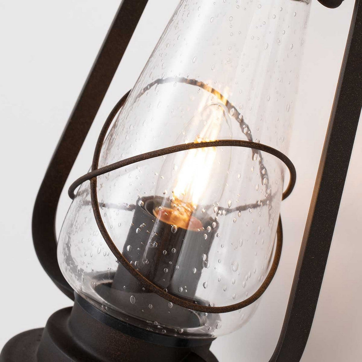 Close-up of the Miners 1 Light Outdoor Wall Light showcasing seeded glass and rustic metal accents in an old bronze finish. The vintage-style lantern casts a warm, glowing light, with raindrop-patterned glass adding to its charm.