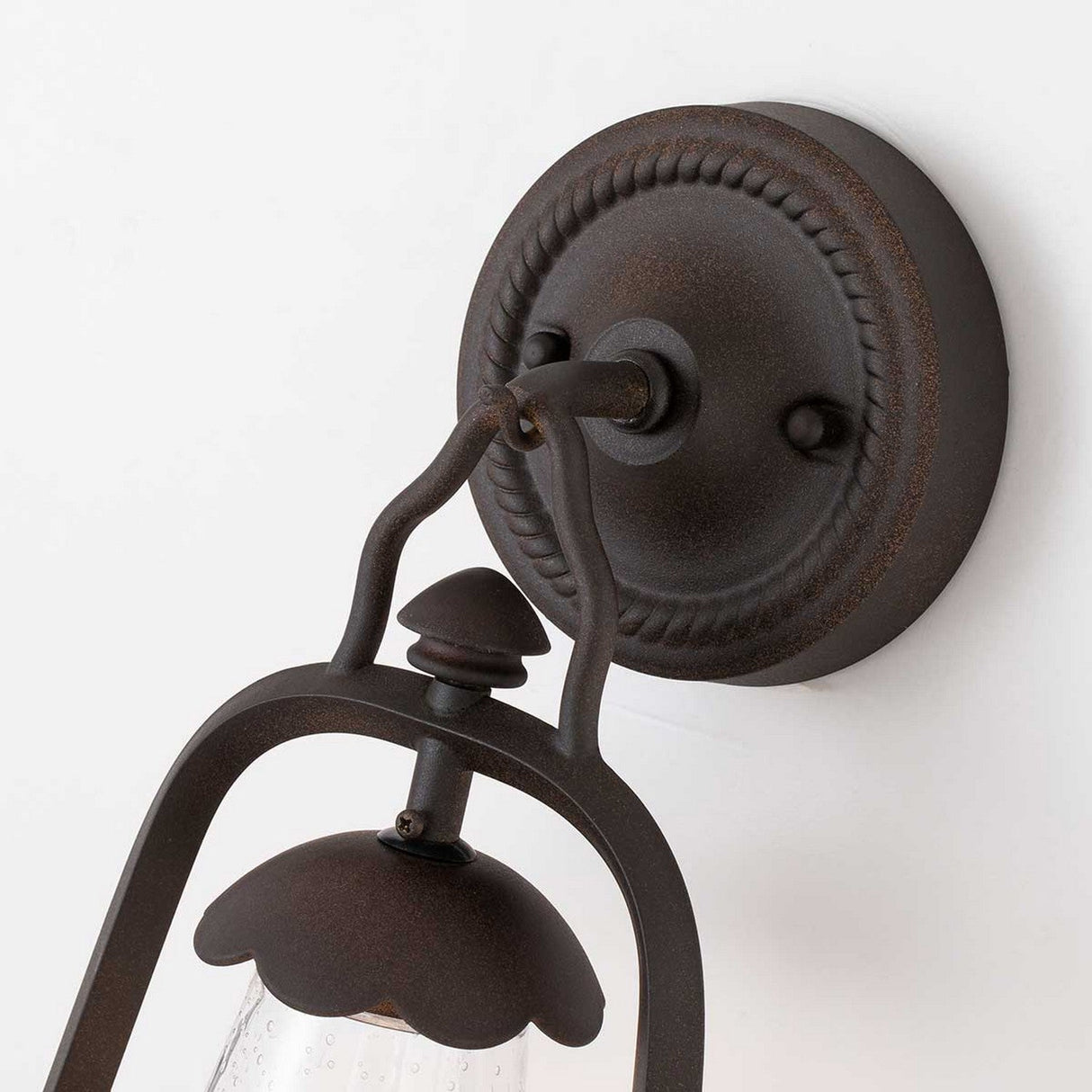 A close-up of the Miners 1 Light Outdoor Wall Light in an Old Bronze finish showcases its vintage style with a decorative round backplate and intricate metalwork. Subtle seeded glass accents are highlighted as the fixture is elegantly mounted on a white wall.