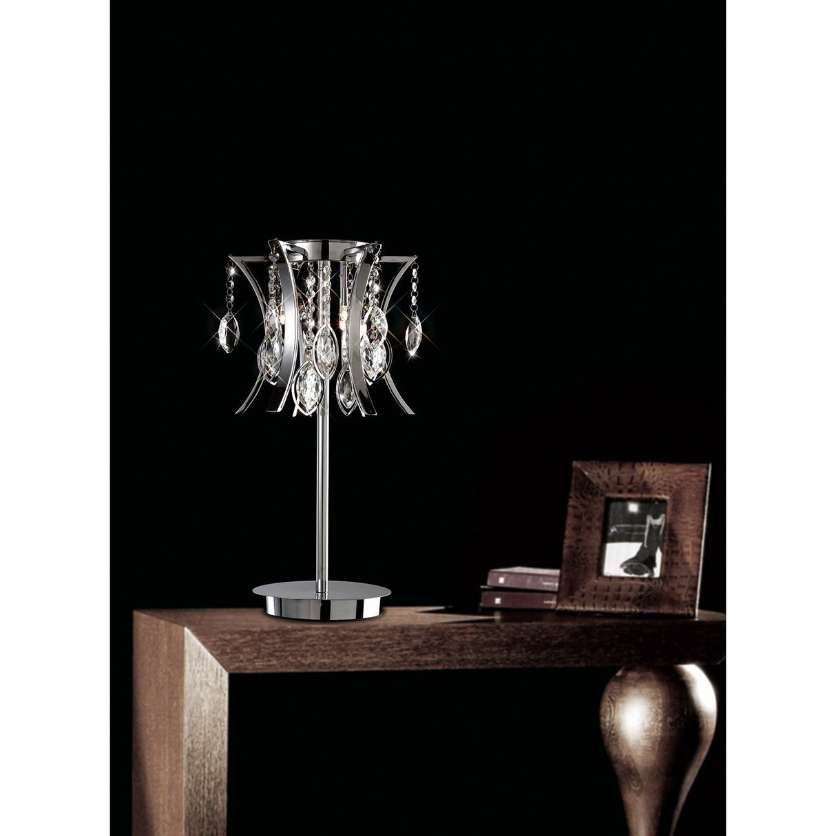 The Mios 3 Light Crystal Table Lamp in polished chrome sits on a wooden table beside a small dark picture frame and a closed book, all set against a solid black background.