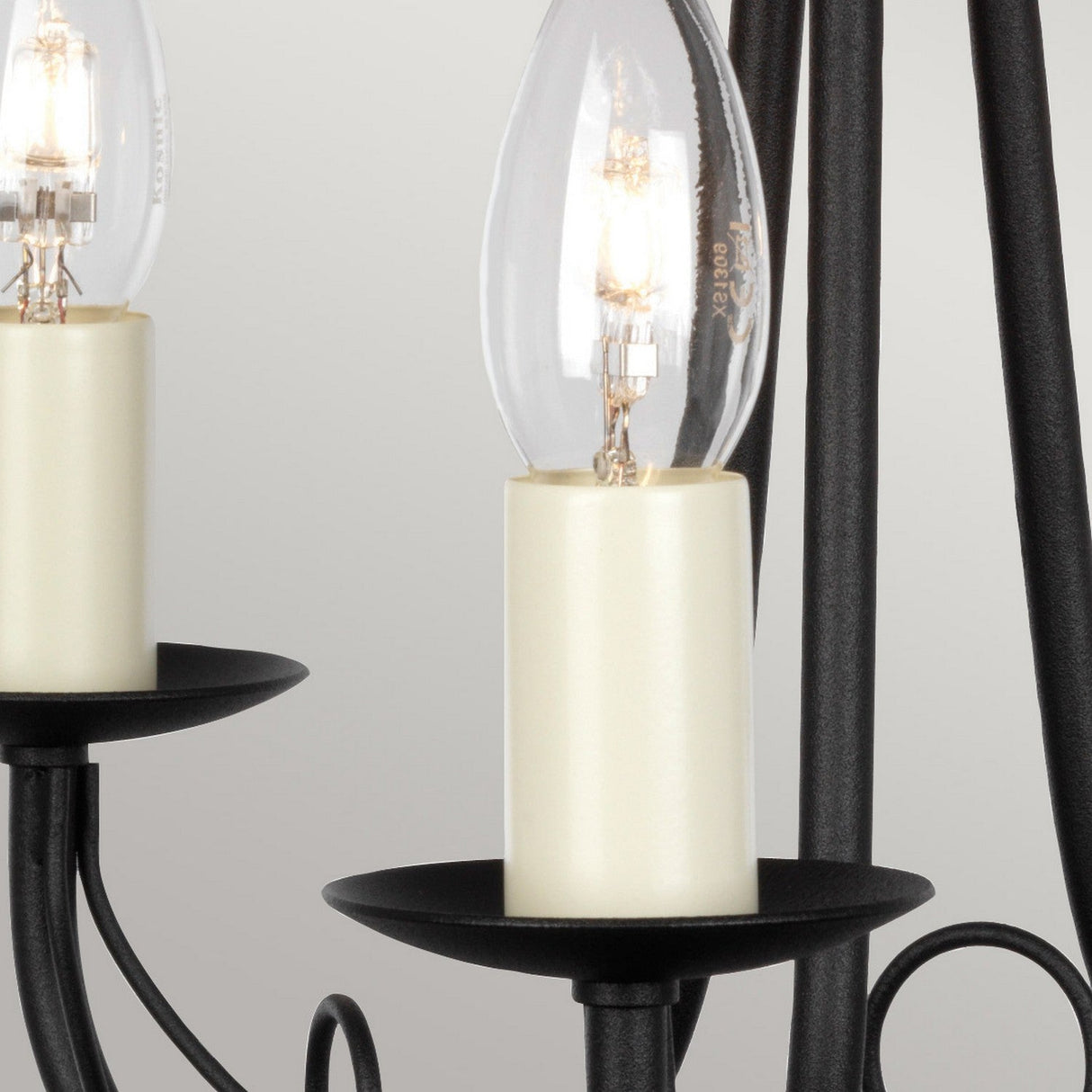 A close-up view features two candle-style light bulbs on the Minster 3 Light Chandelier - Black. The bulbs rest in cream-colored holders with softly glowing filaments, framed by rods and decorative loops of the semi-flush fitting in the background.