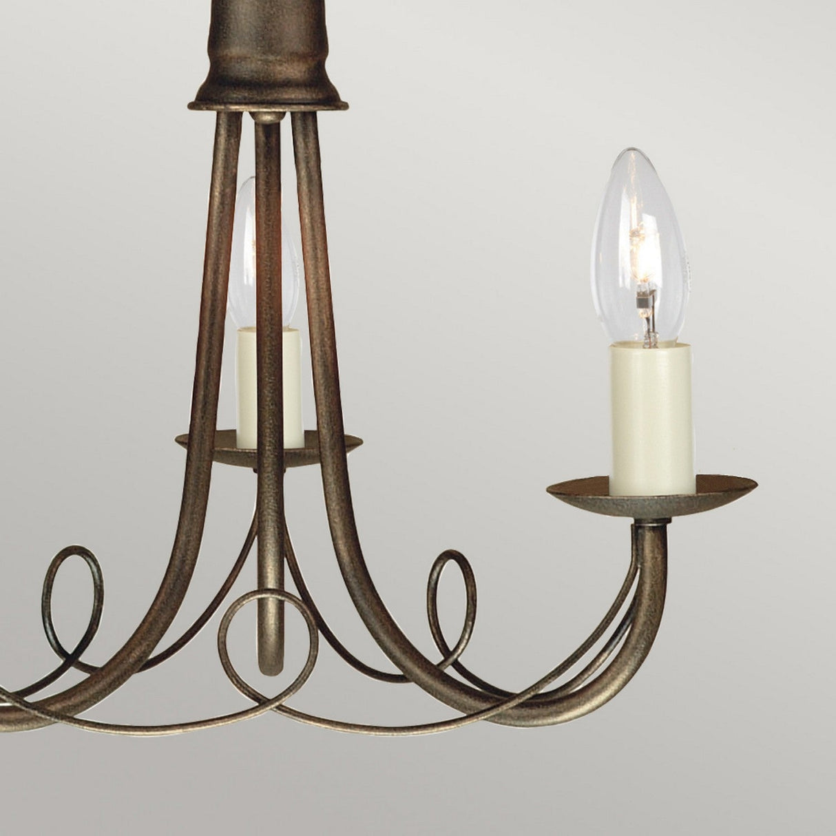A close-up of the Minster 3 Light Chandelier in black with a gold patina, showcasing its ornate design with two candle-like bulb holders. The metal frame features elegant curved and looped patterns. The bulbs resemble flame-tip candles, adding a vintage charm to this semi-flush fitting fixture.