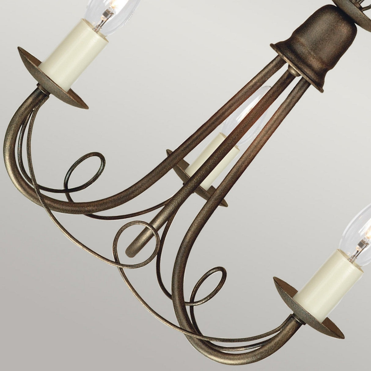 A close-up of the Minster 3 Light Chandelier in black with gold patina highlights its elegant design, featuring three curving arms, each adorned with a candle-shaped light bulb. The semi-flush fitting is accented by scroll-like details and cream-colored bulb bases with a distressed gold finish, set against a light gray background.