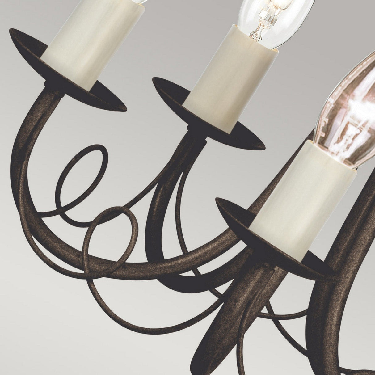 A close-up of the Minster 5 Light Chandelier - Black/Gold Patina highlights its elegant finish, characterized by curved dark metal arms and candle-shaped lights. The convertible semi-flush fitting is striking against a simple, soft gray backdrop.