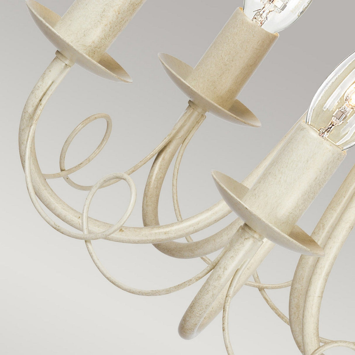 A close-up of the Minster 5 Light Chandelier - Ivory/ Gold Patina reveals its elegantly curved metal arms and exposed bulbs. The metal features a distressed ivory finish accented with gold patina, giving it an antique appearance. Its graceful design is highlighted against a neutral gray background.