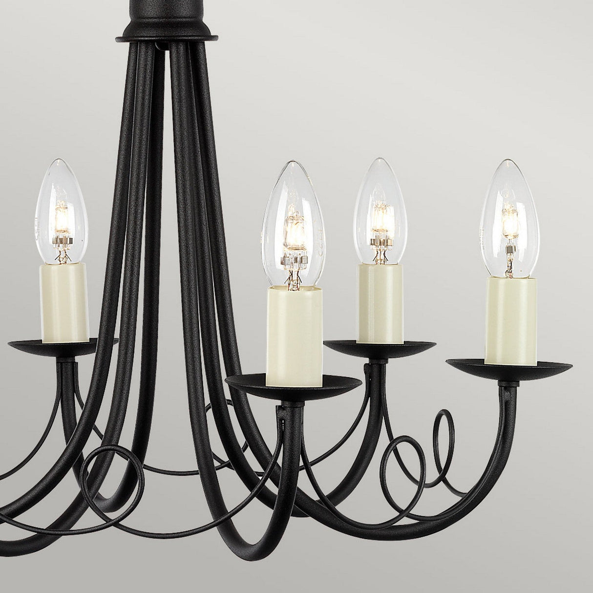 A close-up of the black Minster 6 Light Chandelier highlights its adaptable lighting, featuring six candle-shaped, lit bulb lights. It showcases elegant curving arms and a simple, classic design against a plain gray background.