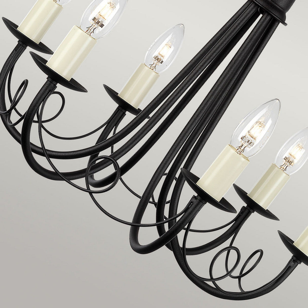 A close-up of the elegant Minster 6 Light Chandelier - Black showcases its black iron finish and six candle-style light bulbs. The gracefully curved arms form a symmetrical pattern, offering a perfect blend of modern and classic design against a neutral background.