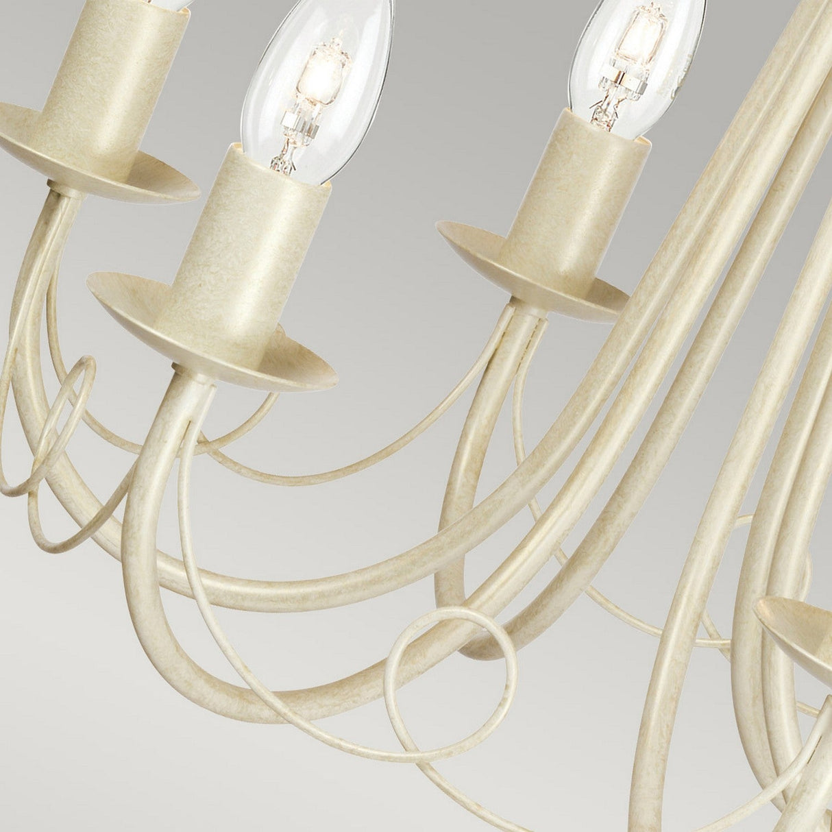 Close-up of the Minster 6 Light Chandelier, highlighting its beige curving metal arms and exposed light bulbs against a neutral background. The elegant design boasts an ivory gold patina finish, offering a classic and sophisticated appearance.