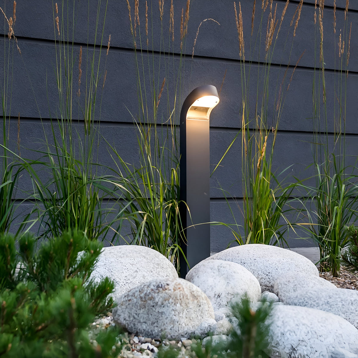 The Molde LED Bollard in Graphite casts a contemporary glow over a garden adorned with tall grasses and smooth white stones. Crafted from die-cast aluminum, it stands elegantly against a dark paneled wall, enhancing the serene ambiance.