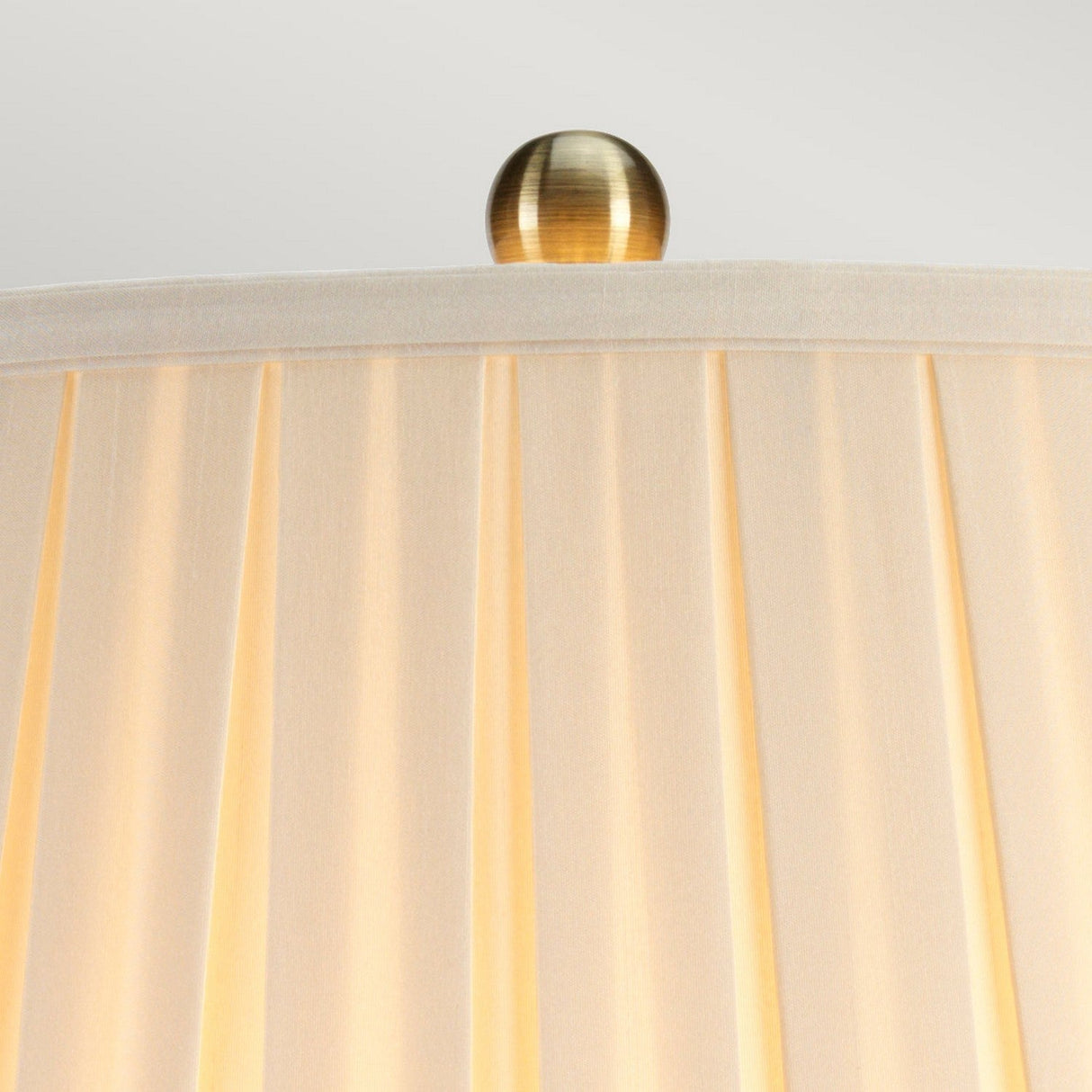 A close-up of the top portion of a beige fabric lampshade with gentle pleats, topped with a small, smooth, metallic finial. The soft lighting highlights the texture of the pleated fabric and subtly complements the elegant Morris 1 Light Large Table Lamp in Gold & Black beneath it.