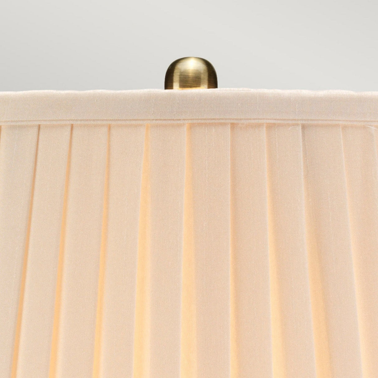 Close-up of a beige pleated lampshade with a brass finial on top, sitting elegantly on the Morris 1 Light Small Table Lamp in gold and black. The pleats create a textured pattern against the backdrop of a plain light gray.