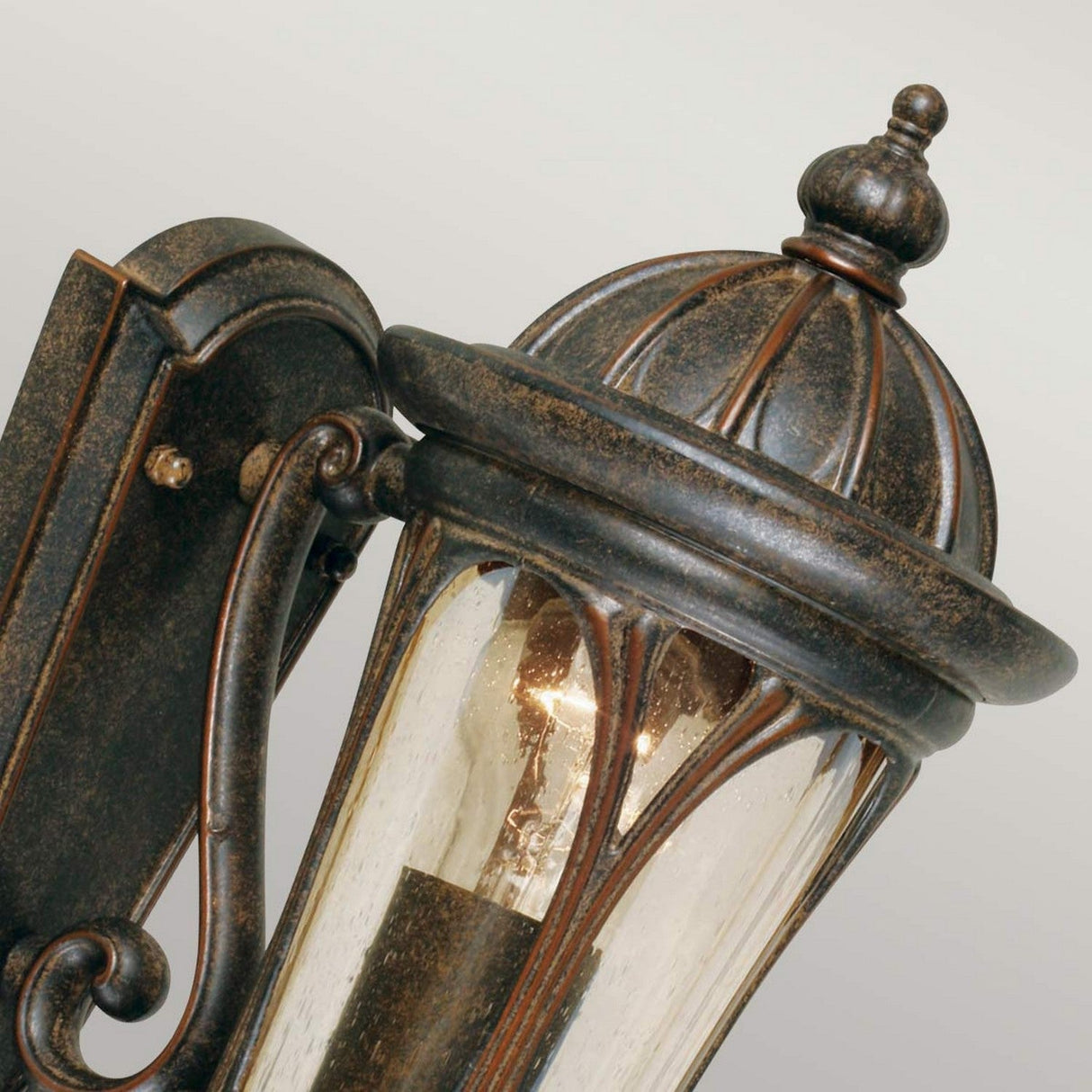 A close-up of the New England 1 Light Outdoor Wall Light showcases its vintage-style and ornate design. The lantern features a weathered bronze finish and clear hand-blown glass panels, revealing an exposed light bulb within. The background displays a soft gradient.
