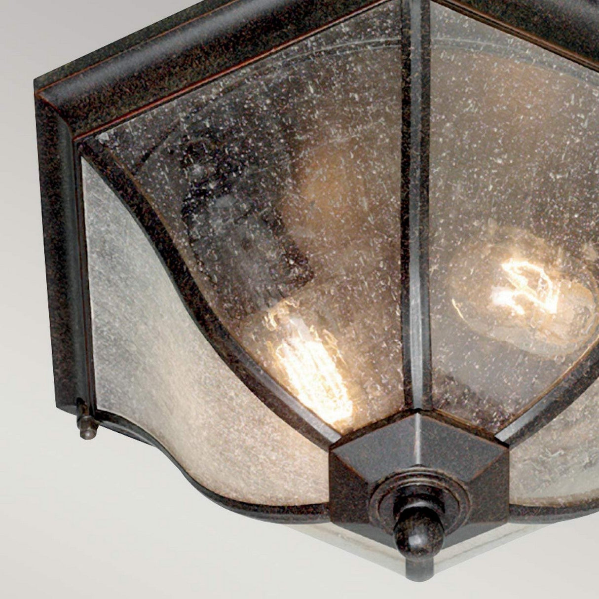 The New England 2 Light Outdoor Flush Light in weathered bronze showcases a vintage appeal with its hexagonal shape and dark metal frame. It features two exposed light bulbs inside, and the glass has a textured, speckled appearance.