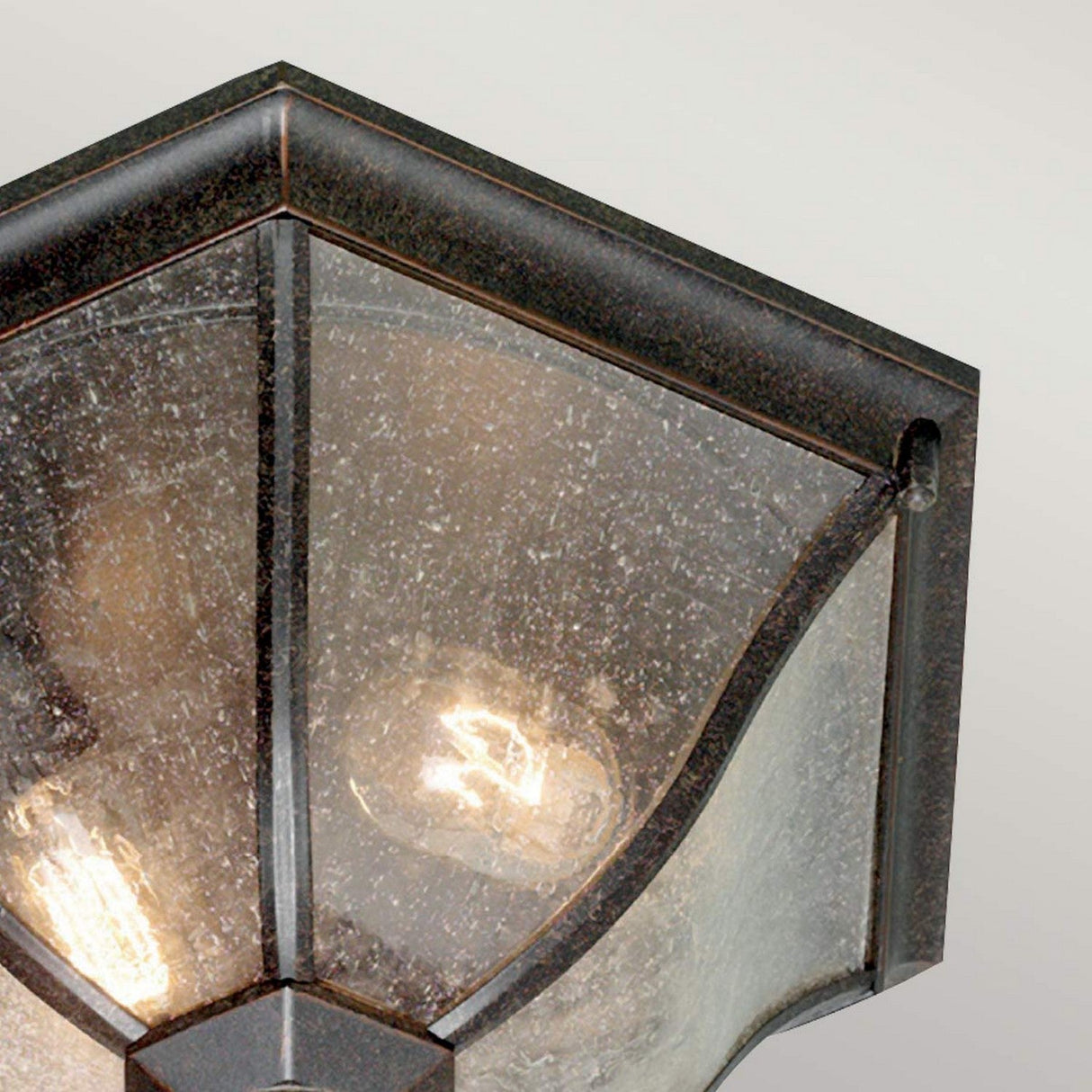 A close-up of the New England 2 Light Outdoor Flush Light in a weathered bronze finish featuring speckled glass panels, through which two light bulbs cast a warm glow that enhances its vintage appeal.