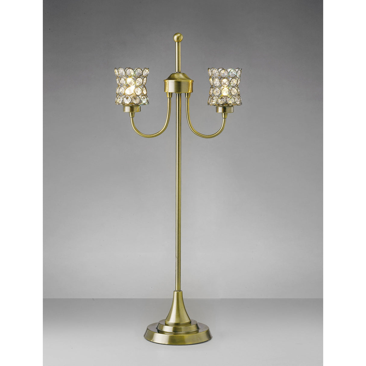 The Nelson 2 Light Crystal Table Lamp in antique brass features two arms, crystal-embellished shades, a round base, vertical stem, and decorative finial. It's shown against a plain gray background.