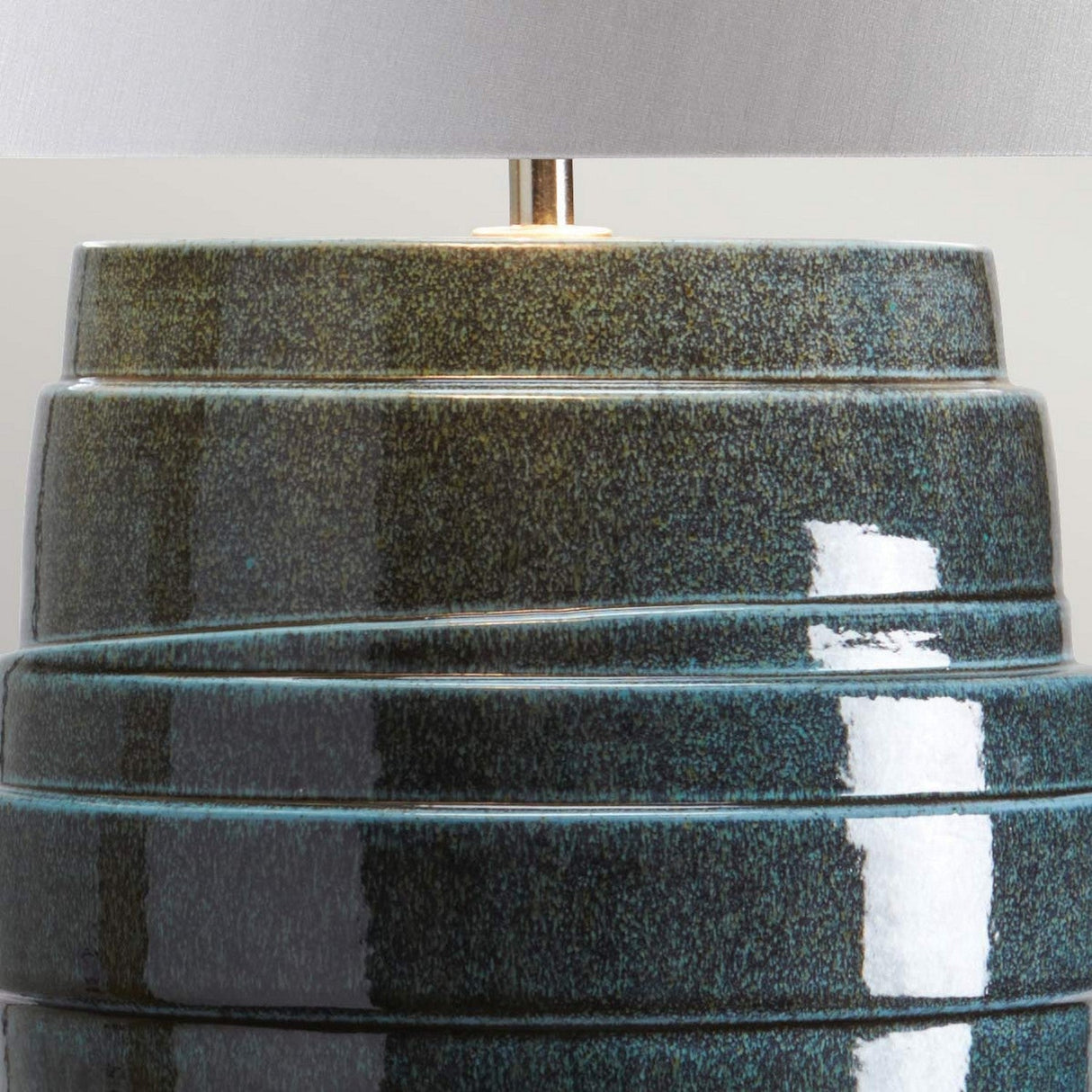 A close-up view of the textured, dark green ceramic base features a tiered, spiral design complemented by polished nickel accents. The base reflects light, showcasing its glossy finish. Partially visible above is a soft gray fabric lampshade that completes the elegant look of the Neptune 1 Light Table Lamp - Blue Glaze.