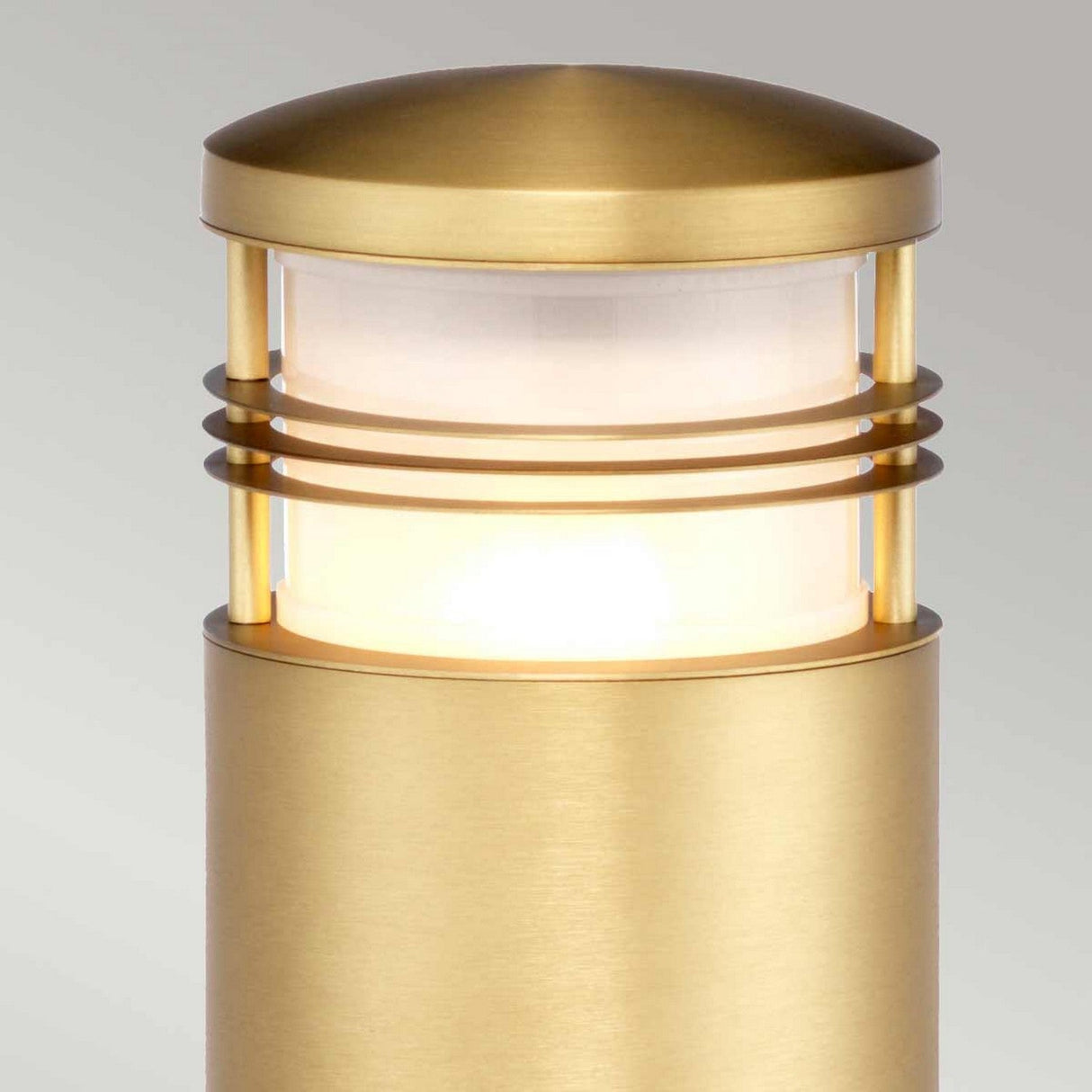 The Newbrighton 1 Light Bollard in brushed brass is an elegant outdoor light fixture with a cylindrical shape. It features a frosted glass section encircled by three horizontal metal bands, which emits a soft, warm glow.