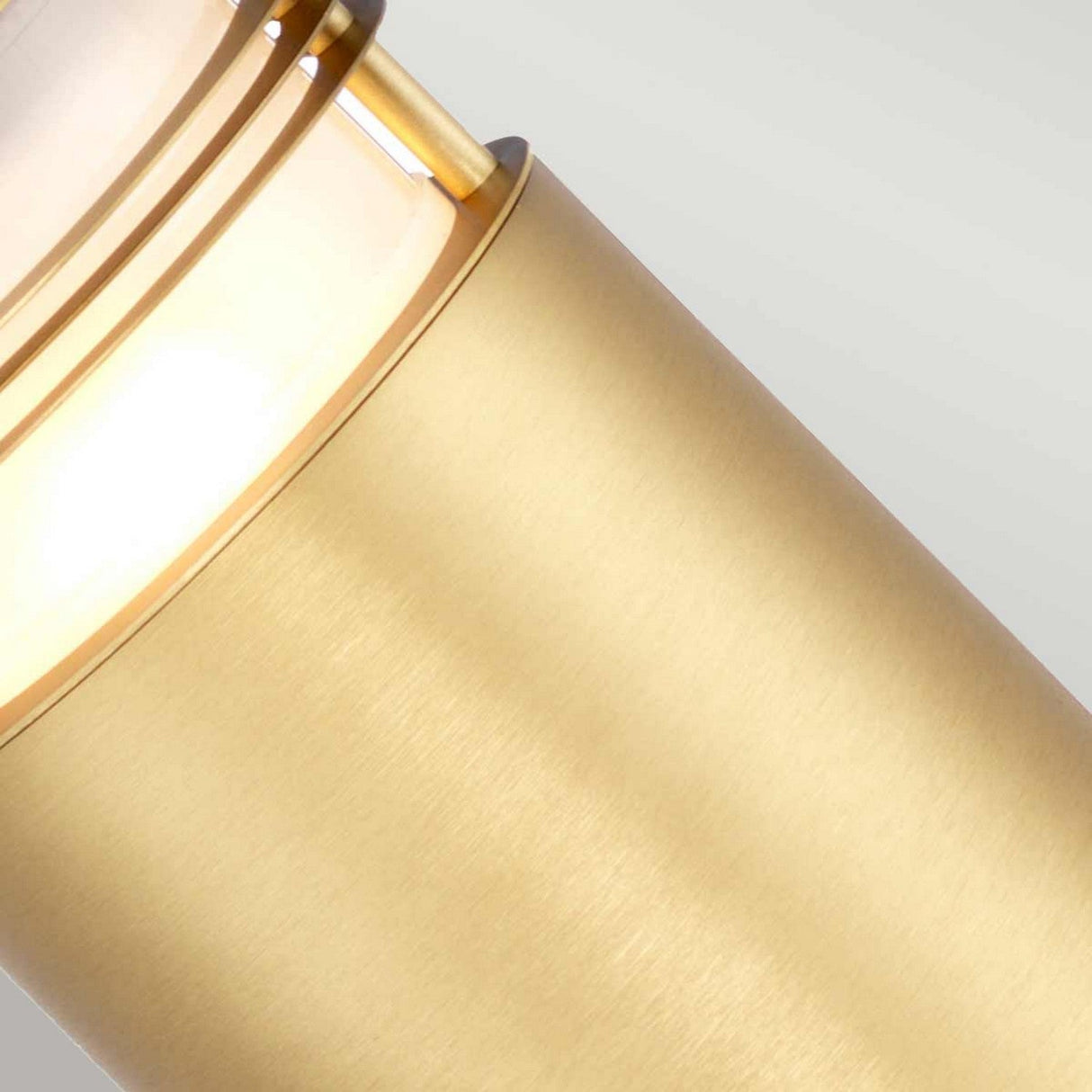 Close-up of the Newbrighton 1 Light Bollard in brushed brass, emitting a soft, warm glow. The cylindrical fixture features a smooth, polished metal surface and a partially diffused light through a frosted glass section. Ideal for outdoor lighting, it is set against a neutral light gray background.