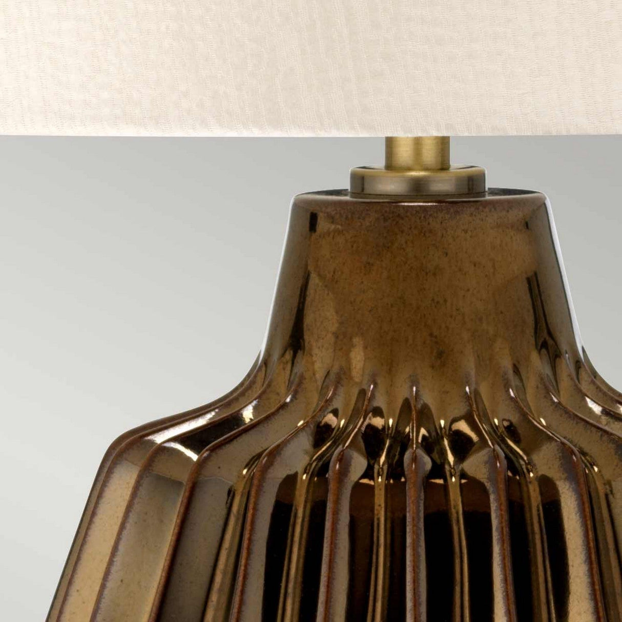 The Newham 1 Light Small Table Lamp in Bronze Ceramic features a close-up of its reflective brown glazed base with vertical grooves. It is topped with a pearl-textured satin shade and brass fitting, set against a soft gray background.