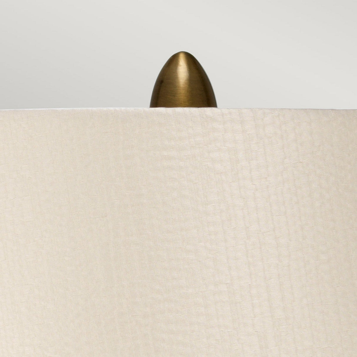 Close-up of the Newham 1 Light Small Table Lamp, showcasing its pearl fabric lampshade and metallic finial. The lamp features a bronze ceramic base, adding elegance against the soft, light gray background.