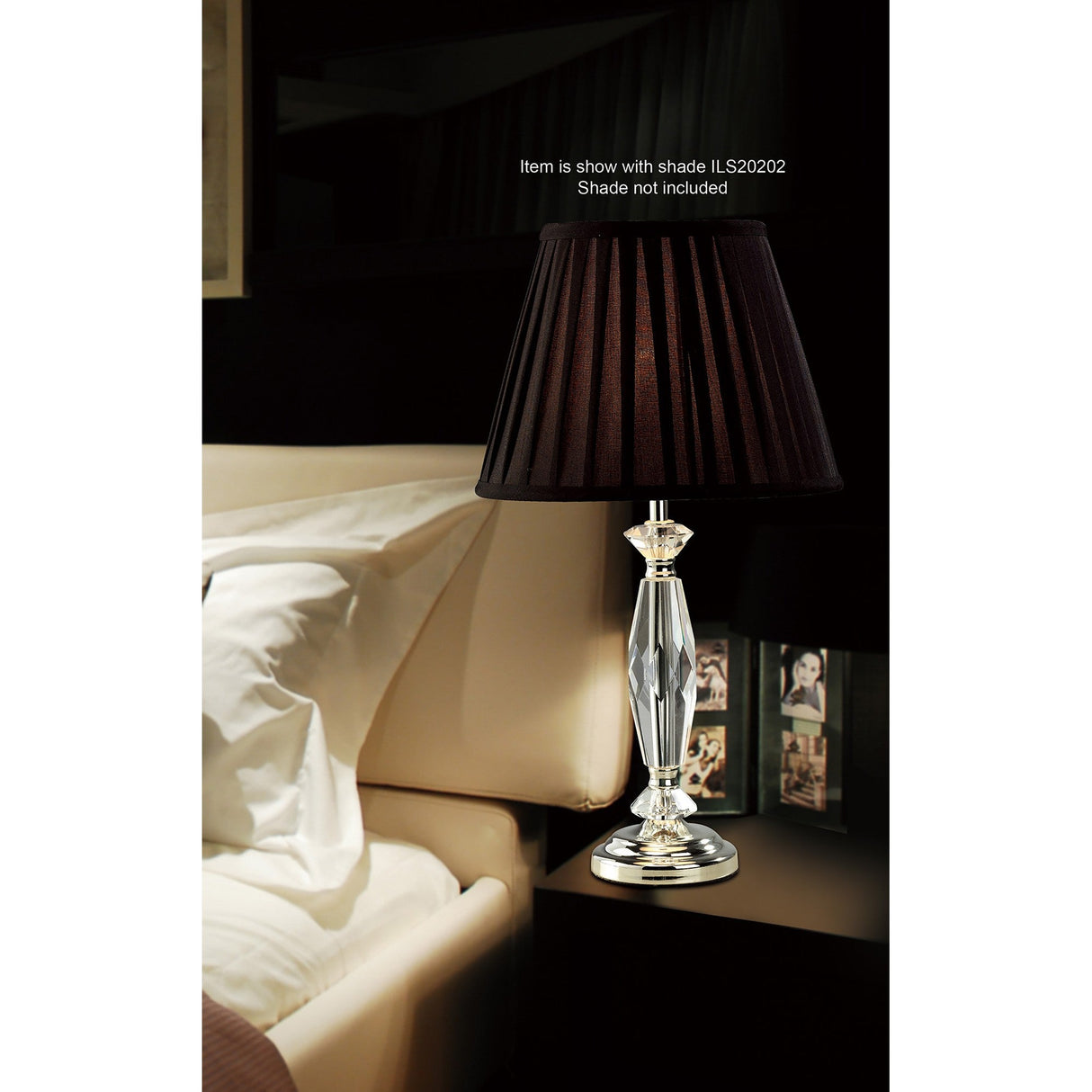 The Nexon 1 Light Crystal Table Lamp in a silver finish, featuring a clear faceted base and black pleated shade, sits on a bedside table beside a beige upholstered bed with framed photos visible in the background.