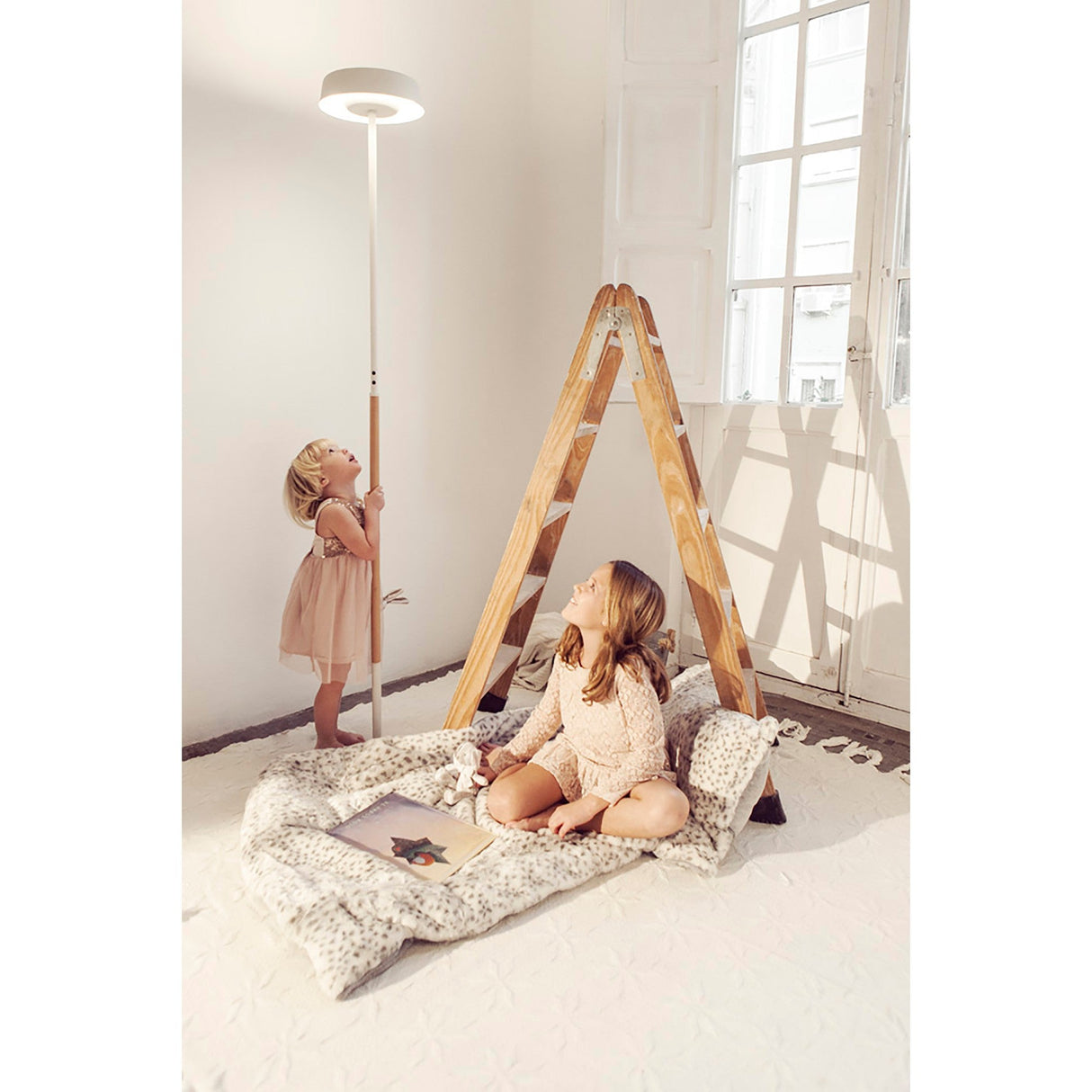 Two young girls in light dresses play indoors. One sits under a wooden teepee, while the other stands by the Noa II Touch Dimmer LED Floor Lamp 9W 3000K - Matt White & Beech. Soft, warm light fills the room.