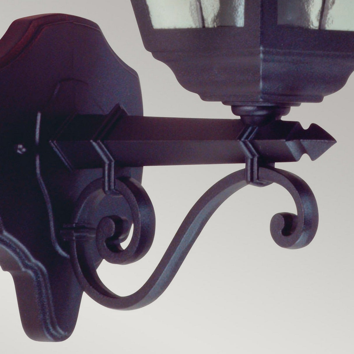 A close-up of the Norfolk 1 Light Up Outdoor Wall Light in black displays its decorative metal design, featuring intricate scrollwork and hand-leaded glass. Mounted on a wall, it combines elegant curves and straight lines to exude a traditional lantern appearance.