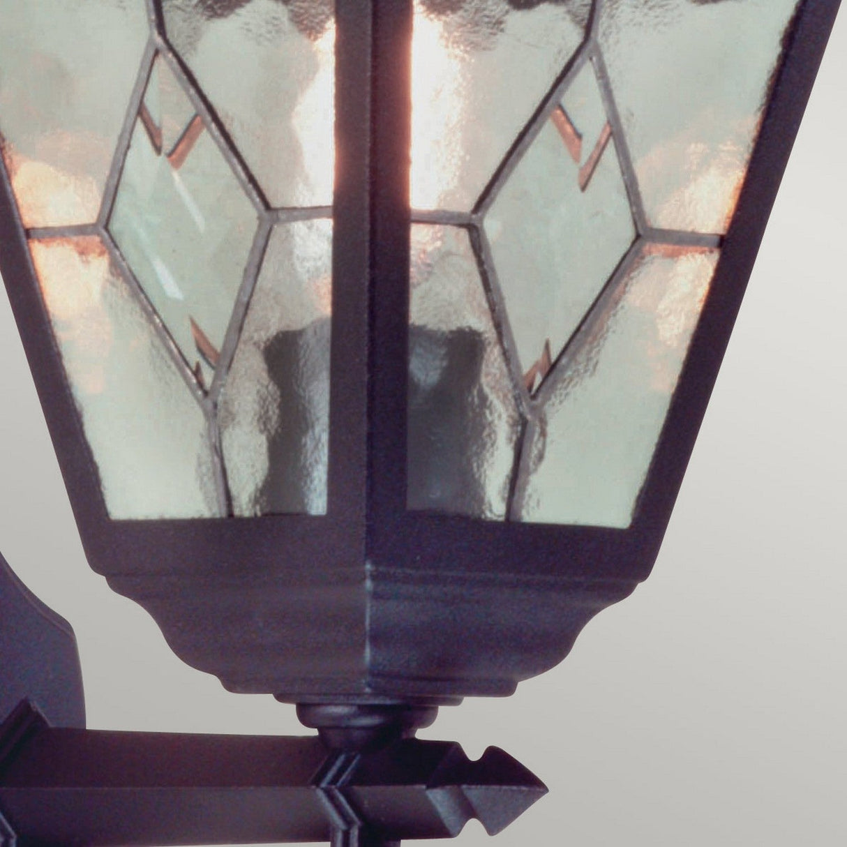 Close-up of the Norfolk 1 Light Up Outdoor Wall Light - Black, showcasing its traditional lantern design. The black frame encases hand-leaded glass panels adorned with a geometric pattern. When lit, it casts a soft glow through the textured glass.