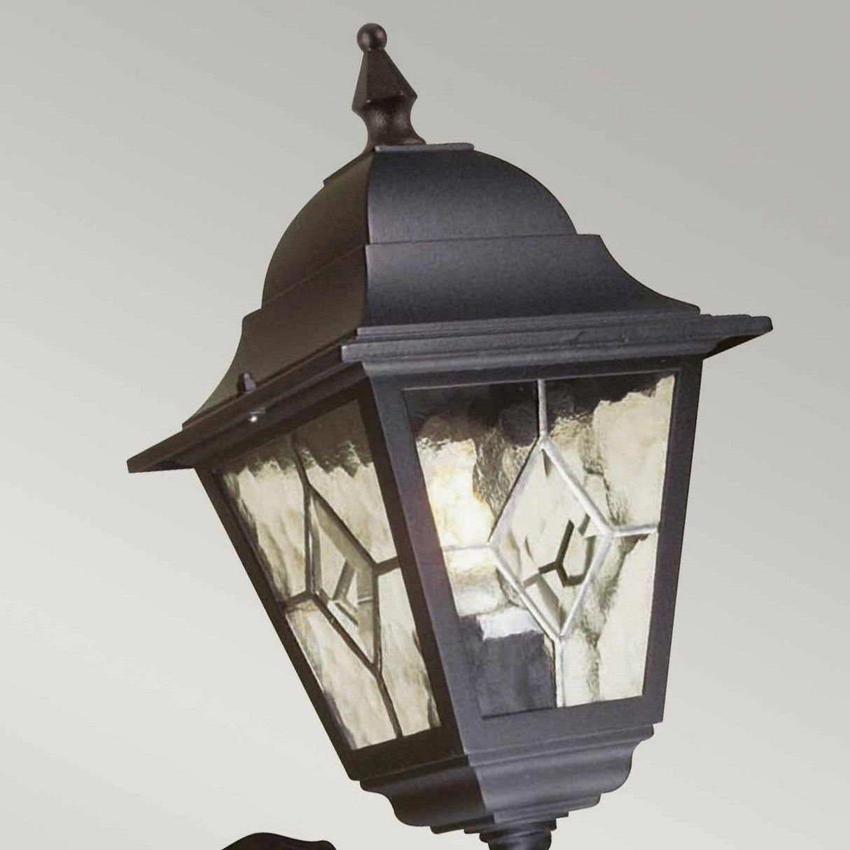 A vintage-inspired outdoor wall light in black, the Norfolk 1 Light Up Outdoor Wall Light with PIR features a pointed top and decorative glass panels adorned with diamond designs. Mounted on a pole, it offers both durability and a slightly weathered appearance suitable for outdoor use.