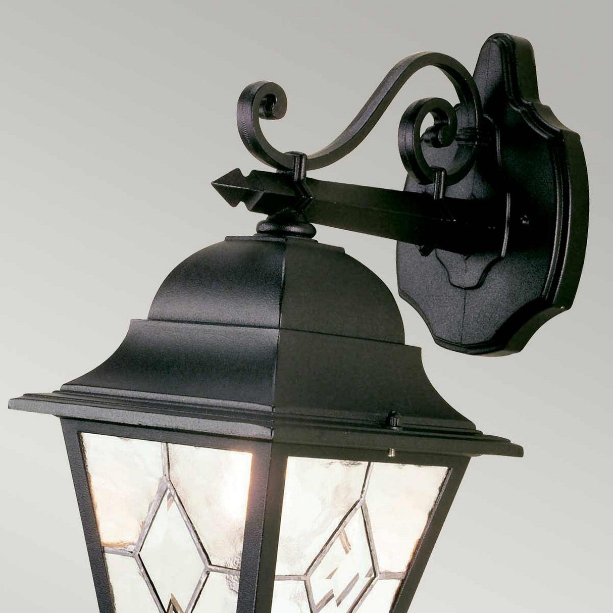 The Norfolk 1 Light Outdoor Down Wall Light - Black features a traditional lantern design with a decorative scroll arm and frosted hand-leaded glass panels showcasing geometric patterns. It is mounted on a wall bracket with a smooth, matte finish.