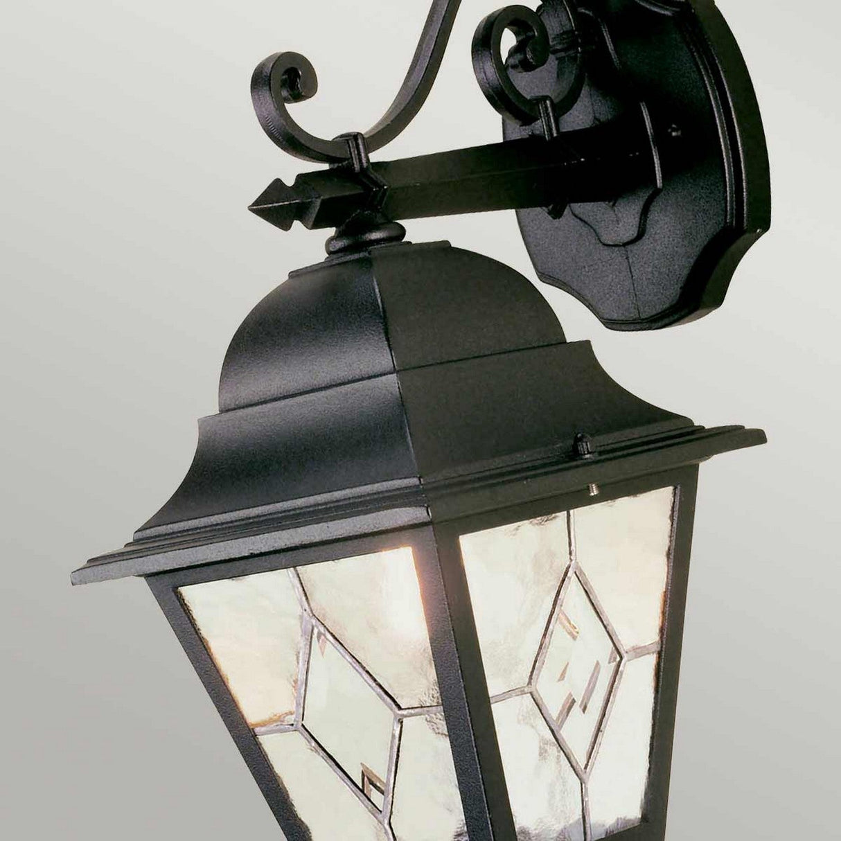 The Norfolk 1 Light Outdoor Down Wall Light in black boasts a traditional design, adorned with hand-leaded glass panels featuring diamond-shaped patterns. This classic lantern is elegantly mounted on a curved bracket against a light gray background.