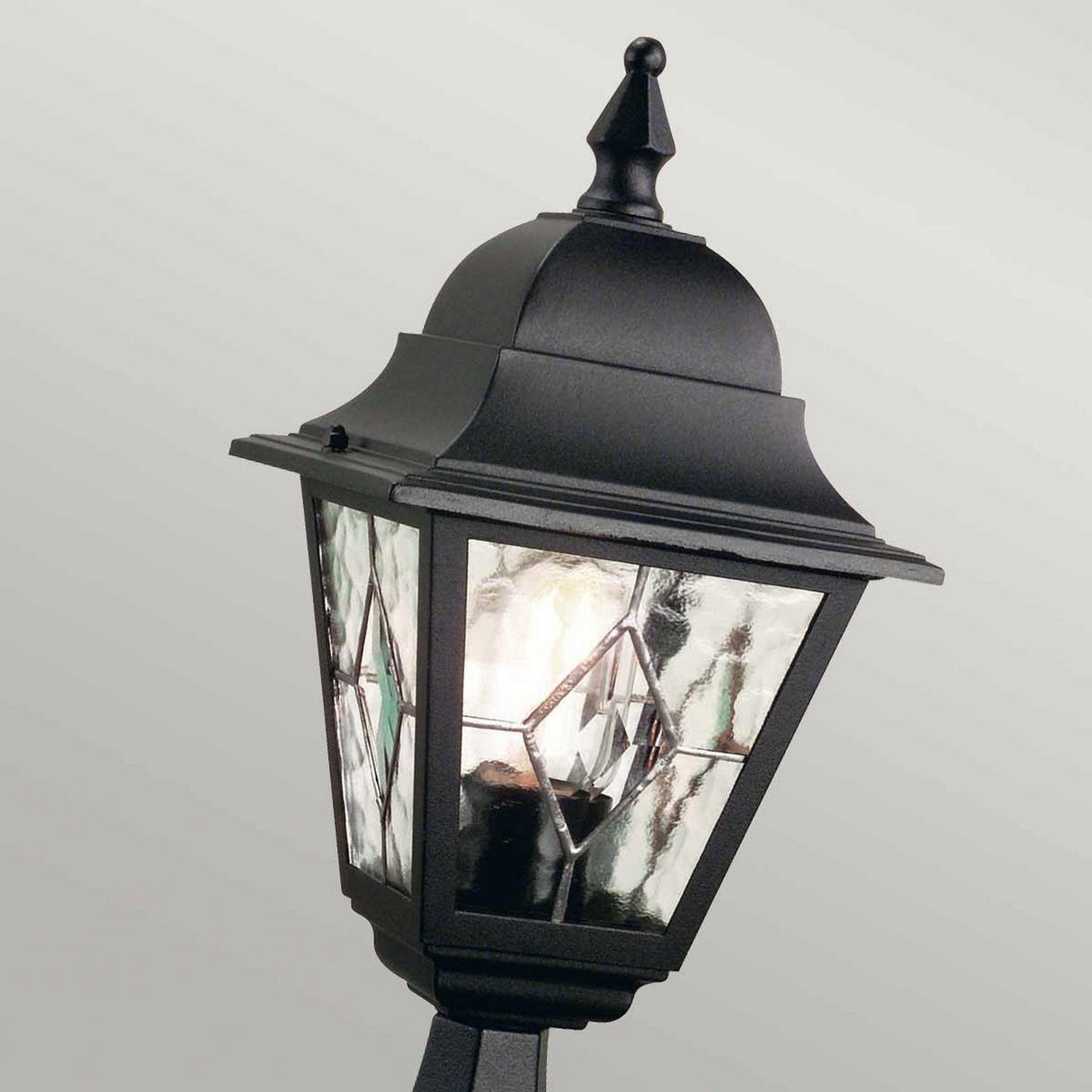 The Norfolk 1 Light Pedestal Light - Black is an elegantly designed outdoor lantern featuring hand-leaded, frosted glass panes and a pointed top. Mounted on a post at a slight angle, it emits a soft glow that beautifully highlights the intricate patterns of the glass.