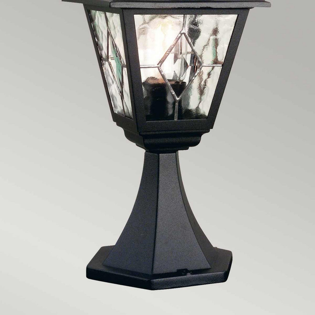 Close-up of the Norfolk 1 Light Pedestal Light in black, featuring a decorative outdoor lantern with a metal base and hand-leaded glass panels that emit a soft light. This fixture showcases a classic, elegant design that elevates any space with its sophisticated outdoor lighting.